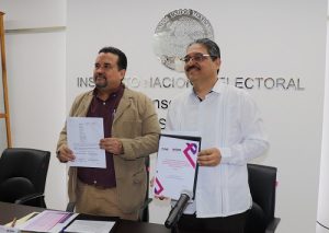 Dos hombres, uno de lente, de pie posando con convenio firmado