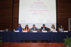 Integrantes del presídium del evento sentados detrás de mesa sobre escenario
