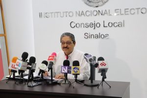 Vocal del INE Sinaloa sentado detrás de mesa con varios micrófonos