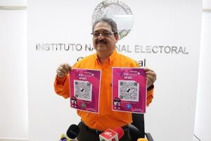 Vocal del INE sosteniendo dos hojas con códigos QR para acceder a la plataforma "Conóceles"