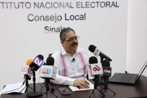 Hombre de lentes y bigote sentado con varios micrófonos frente a él