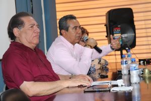 Hombre de camisa rosa claro hablando con micrófono en mano derecha