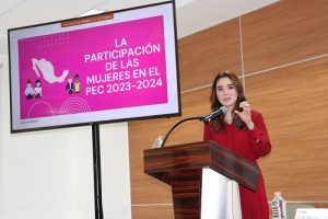 Mujer con vestido rojo hablando en un pódium, atrás un letrero rosa con letras blancas
