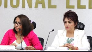 Mujer de lentes junto a mujer de cabello corto, ambas sentadas detrás de mesa