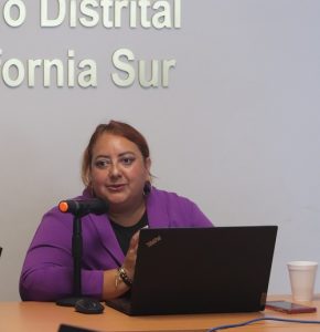 Mujer de cabello rojizo hablando a micrófono sobre mesa