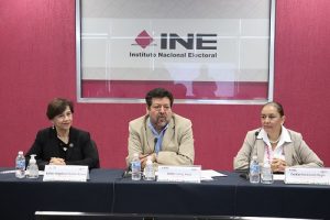 tres personas sentadas frente a una pared rosa con las letras del INE