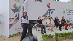 Hombre de pie hablando con micrófono y personas sentadas a un lado