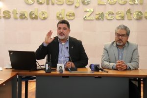 Hombre de barba cana junto a hombre de lentes, cabello y barba cana, ambos sentados detrás de mesa