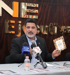 Hombre de barba canosa, sentado detrás de mesa hablando con micrófono en mano