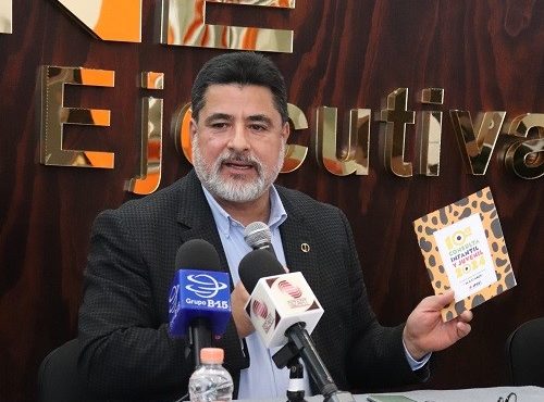 Hombre de barba canosa, sentado detrás de mesa hablando con micrófono en mano