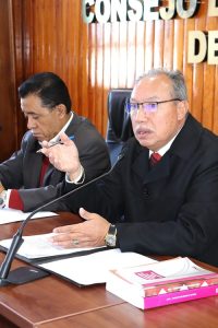 Hombre semicalvo de lentes, sentado detrás de mesa hablando a micrófono