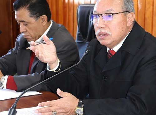 Hombre semicalvo de lentes, sentado detrás de mesa hablando a micrófono
