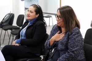 Dos mujeres sentadas, una de ellas usando lentes y con su mano en el pecho