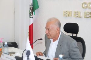 Hombre de cabello cano sentado detrás de escritorio hablando a micrófono