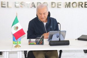 Hombre adulto mayor, sentado frente a una computadora, un micrófono y una mini bandera