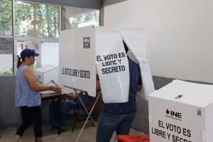 Dos personas de pie, emitiendo su voto, en mampara del INE en Tabasco