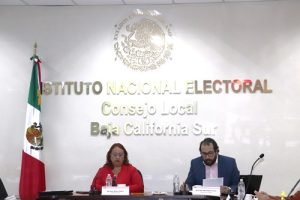 Dos personas sentadas y a un costado está la bandera de México