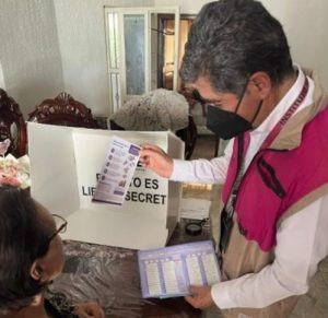 hombre con chaleco del INE entregando boletas a una mujer en el interior de su domicilio
