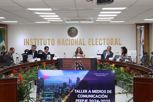 varias personas sentadas alrededor de una mesa rectangular con micrófonos, al frente una pantalla