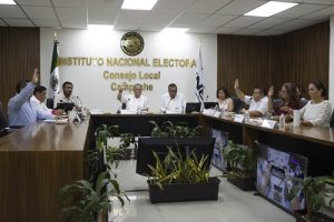 varias personas sentadas alrededor de una mesa rectangular con micrófonos, votando a mano alzada.