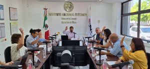varias personas sentadas alrededor de una mesa rectangular con micrófonos, votando a mano alzada