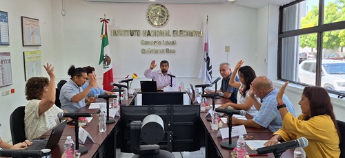 varias personas sentadas alrededor de una mesa rectangular con micrófonos, votando a mano alzada