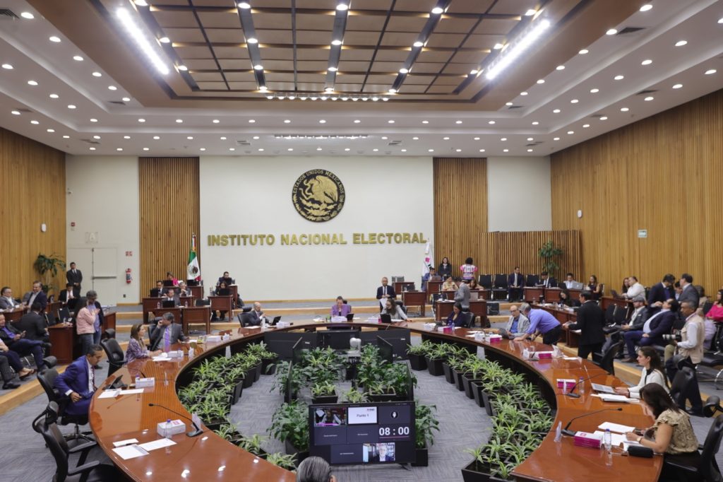 Salón de sesiones con el escudo de México al fondo con letras de color dorado que dice “Instituto Federal Electoral”, mesa en forma de herradura con 10 personas Consejeras Electorales y alrededor de la mesa directores ejecutivos, titulares de unidades técnicas y asesores.
