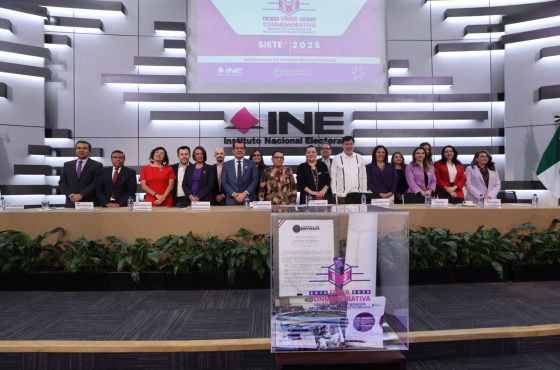 Imagen abierta en el auditorio del instituto con las personas participantes de pie al frente del recinto, pantalla con logo del evento contiene una urna y abajo de ella letrero del Instituto Nacional Electoral.