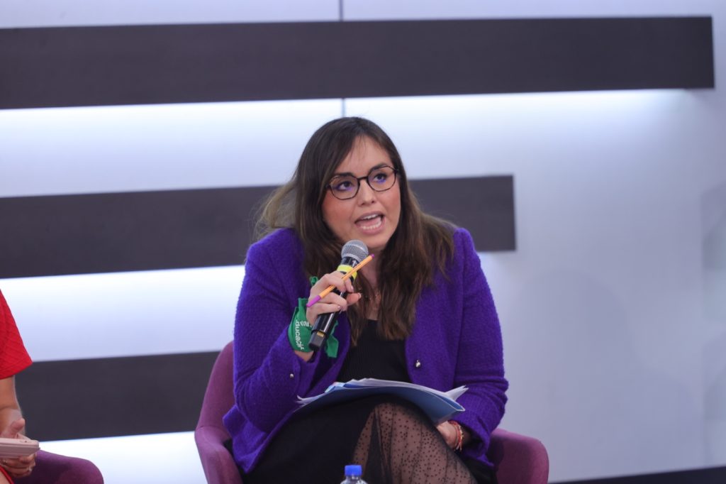Patricia Carranza Rodríguez, Directora General de Inteligencia, Innovación y Planeación estratégica para la mejora continua de los Programas de la Secretaría de las mujeres, sentada hablando en el estrado al frente del recinto.