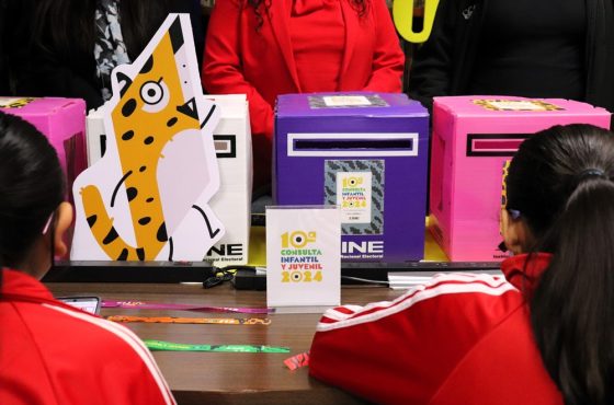 Dos menores de edad sentados viendo cuatro urnas utilizadas en la Consulta Infantil y Juvenil 2024