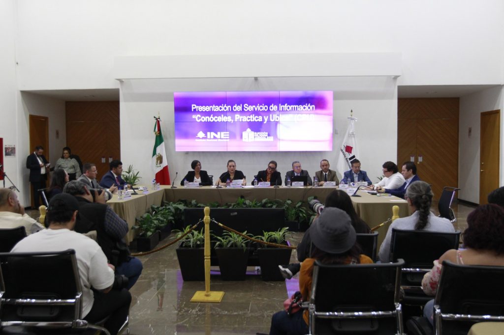 Imagen abierta en el lobby del instituto con las personas participantes sentados en el estrado al frente de la audiencia y letrero del evento.