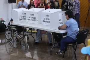 Persona pequeña votando en mampara especial y otra persona votando en mampara especial en silla de ruedas.