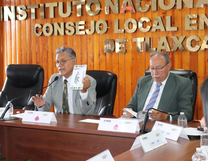 dos hombres sentados, uno de ellos sostiene un cuadernillo blanco con diferentes colores