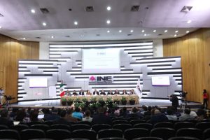 Personas participantes y público asistente al evento sentadas en el lobby del instituto. Personas participantes sentadas en el estrado al frente del recinto.