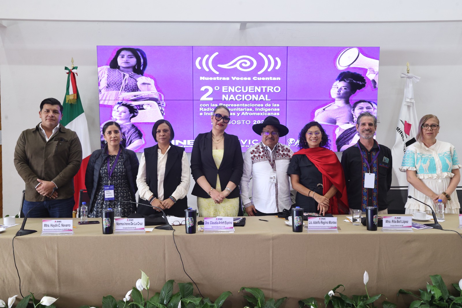 Personas participantes paradas en el estrado al frente del recinto.