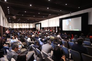 Imagen abierta en el salón del evento con las personas participantes sentadas al frente de la audiencia.