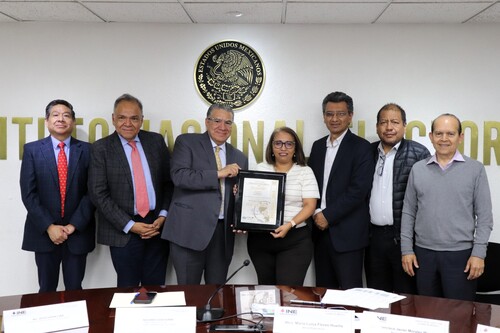 personas de pie frente al escudo nacional mexicano sostienen un documento