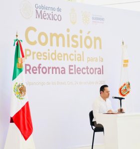 Hombre vestido de blanco sentado frente a un mesa con micrófono