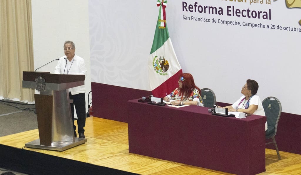 un adulto mayor de pie frente a un atril, a su lado dos mujeres sentadas