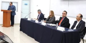 cuatro personas sentadas y uno de pie hablando frente a un micrófono