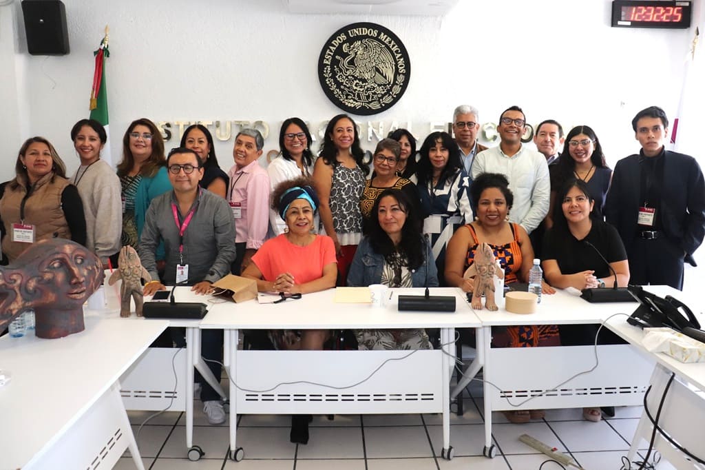 personas sentadas y de pie en un salón blanco con el escudo nacional