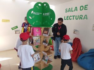 personas menores de edad de pie en un salón con un árbol con libros