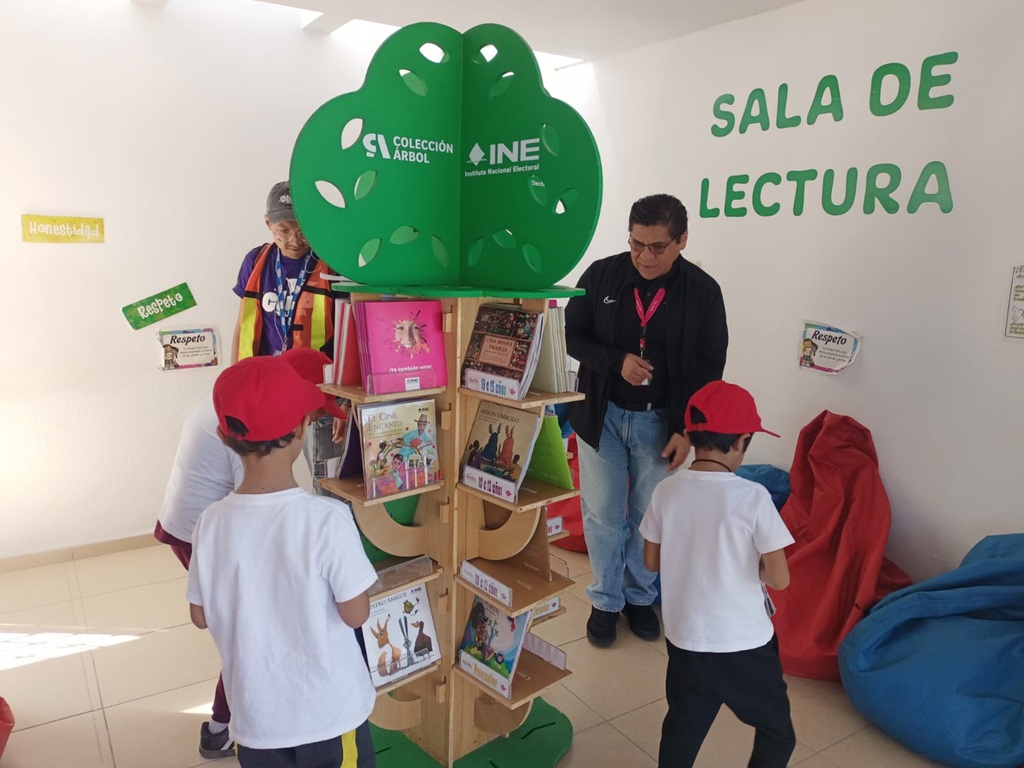 personas menores de edad de pie en un salón con un árbol con libros
