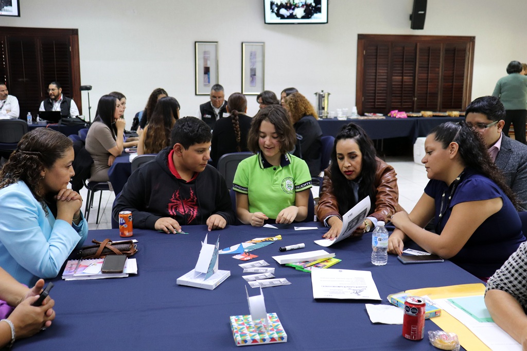 niñas, niños y personas adultas sentadas alrededor de una mesa platicando