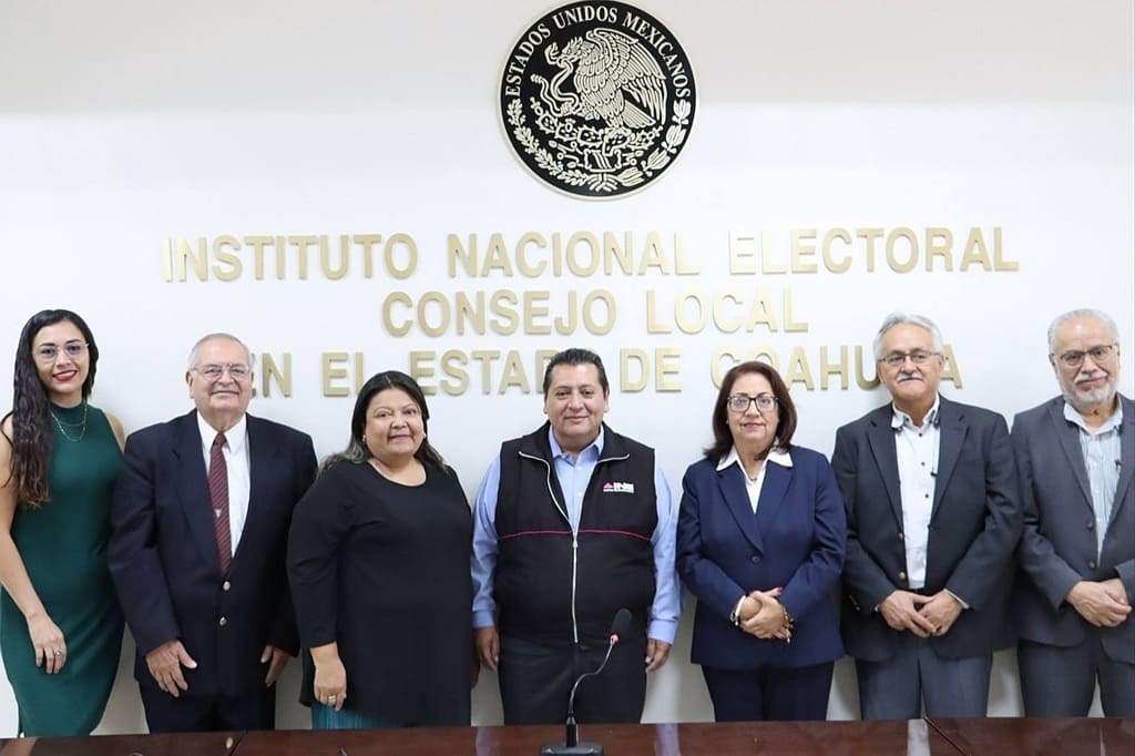 personas de pie najo el escudo nacional vestidas de color oscuro