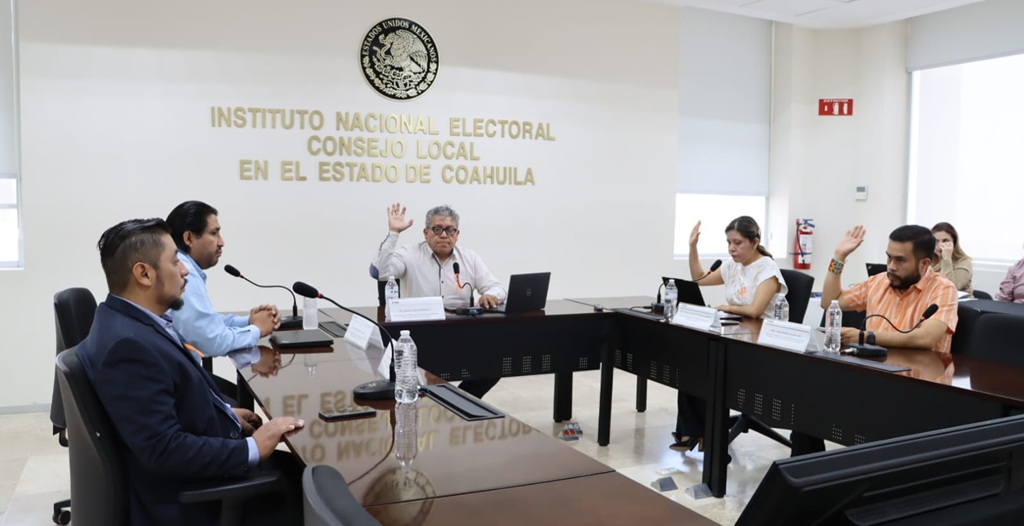 personas sentadas alrededor de una mesa con micrófonos y la mano levantada