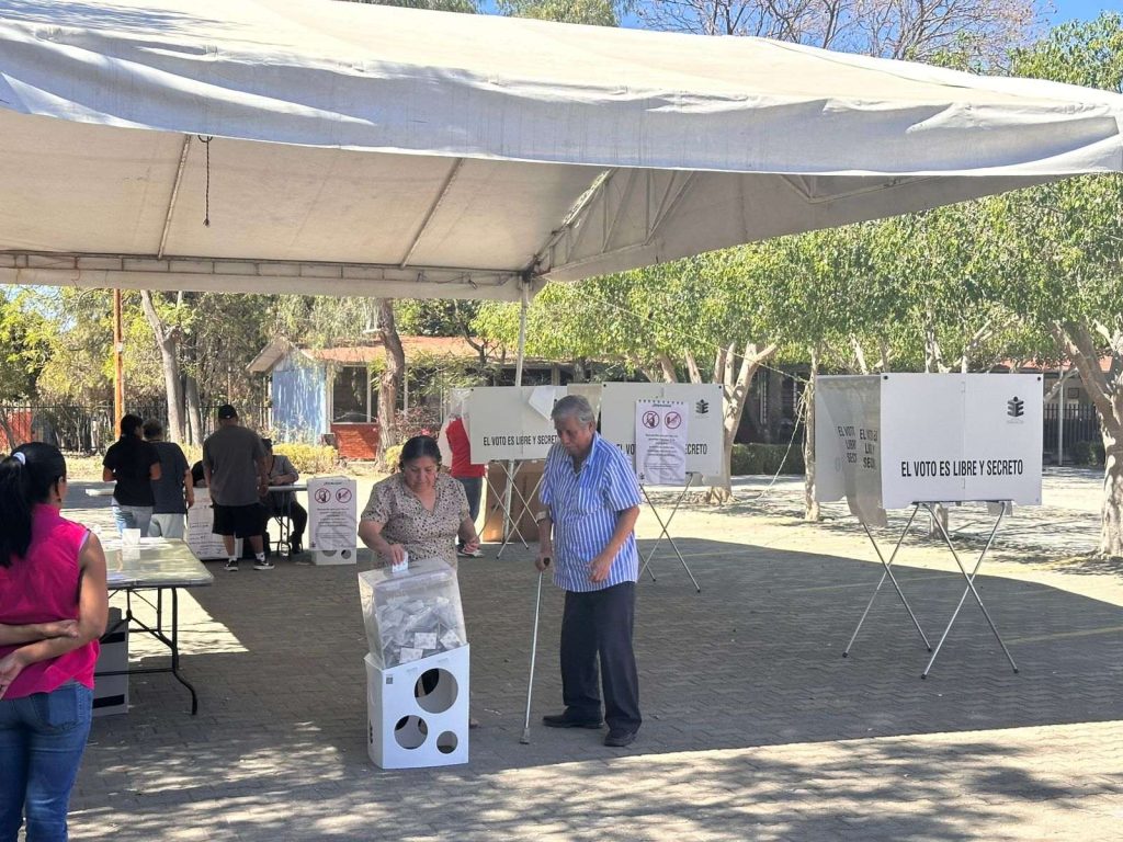 Casilla con personas funcionarias, mampara, urna y personas votando.