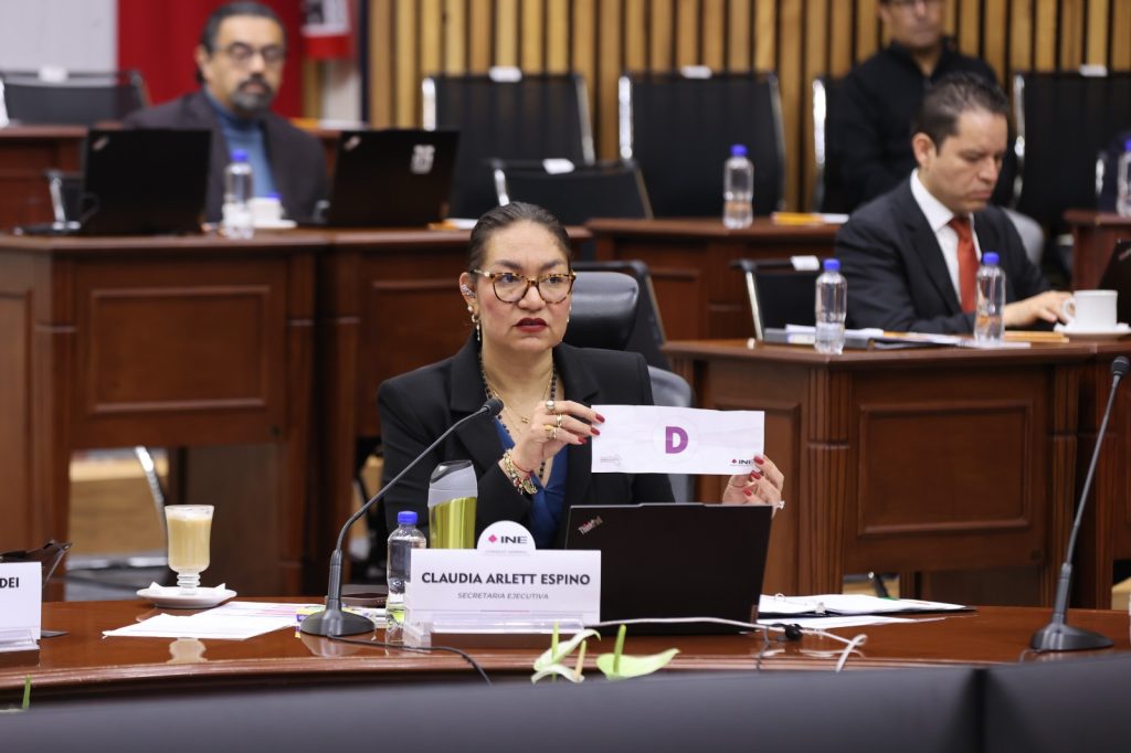 Claudia Arlett Espino, Secretaria Ejecutiva sentada en la herradura mostrando la letra base para seleccionar a la ciudadanía que integrarán las Mesas Directivas de Casilla.