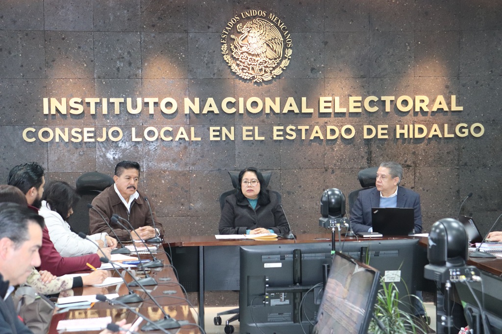 personas sentadas alrededor de una mesa rectangular con una pantalla de Televisión en medio