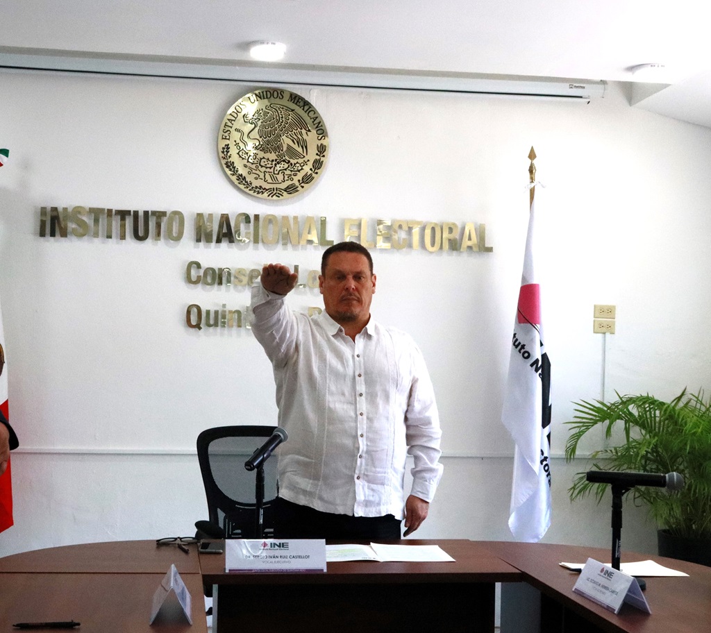 Hombre de pie detrás de una mesa vestido con camisa blanca elevando su brazo derecho en señal de protesta